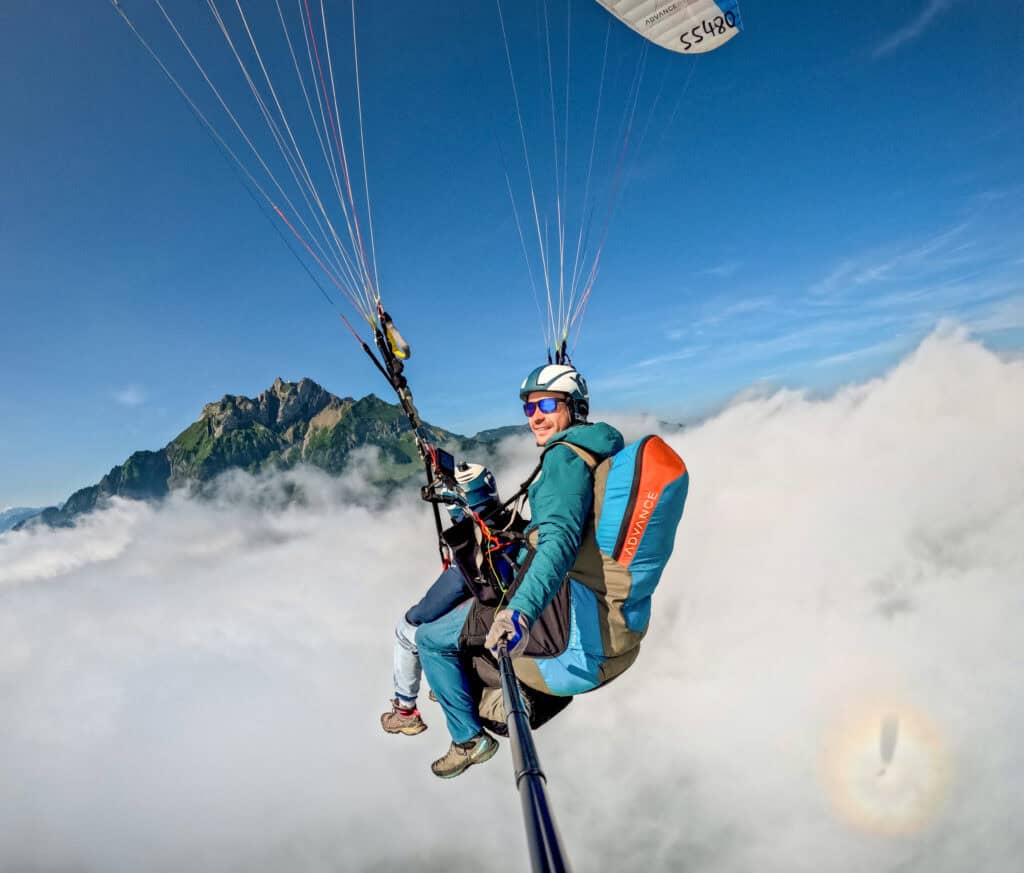 Pilot und Passagier beim Tandemflug betrachten den wolkenverhangenen Pilatus, mit Halo am Himmel und glücklichem Piloten – beeindruckende Stimmung