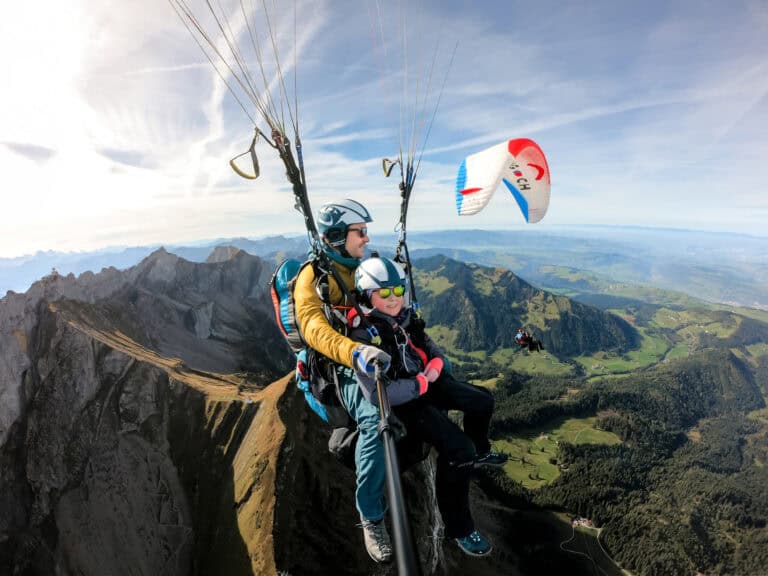 Lächelnder junger Passagier fliegt über der Pilatus-Kette mit leichten Cirruswolken am Himmel; neben ihm ein zweiter Tandemflieger mit seinem Vater, leichter Nebel über dem Schweizer Flachland
