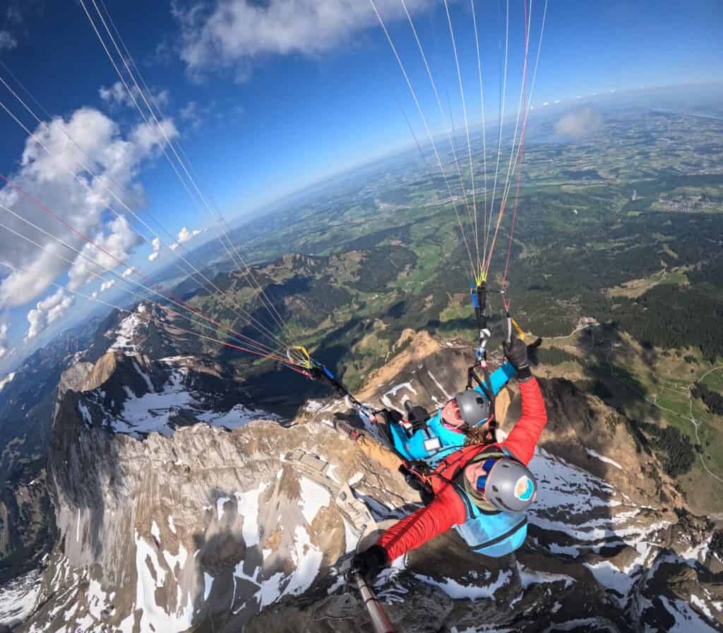 Tandempilot und Passagier schweben unter einem bunten Gleitschirm über dem Pilatusmassiv, mit Blick auf den leuchtend blauen Vierwaldstättersee und die umliegenden Alpen.